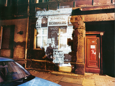 Photograph of blank city wall with projection of storefront over it and car in foreground.