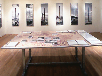 Installation showing set of eight photographs arranged on back wall, and table in foreground with fourteen photographs and fourteen texts lying flat.  