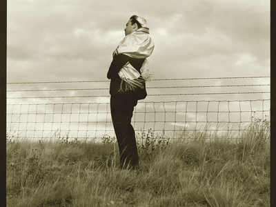 Full body photograph depicting man in kippah and prayer shawl standing in a field next to fence, facing to the side.