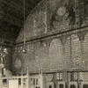 Interior of building of back wall with decorative images on top, Hebrew text in the middle, and lattice below, platform with railing in left foreground, and several podiums along wall.