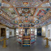 Photograph of room with mural ceiling of floral patterns, animals, and panels of Hebrew text, and painted canopied platform in center of room.  